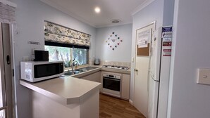 A compact kitchen with a microwave, sink, stove, and oven. There is a window with a view of trees, a clock on the wall, and a door with a bulletin board.
