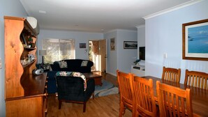 A living room with a dark blue sofa, wooden dining table, and chairs. There is a wooden cabinet with decorative items and a window with blinds.