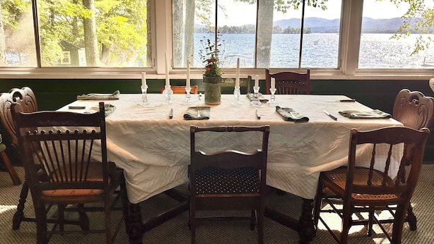 A dining area with a table set for six, large windows with a lake view, and wooden chairs.