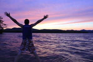 A person standing with arms outstretched in a body of water with a sunset in the background. - Lakefront House & Cottage ~ walk-in beach, hammock, canoe, kayaks and sunsets (Kattskill Bay)