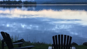 A lakeside view with two Adirondack chairs facing the water, surrounded by mountains and a cloudy sky.