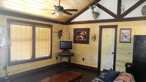 Un interior de madera con ventilador de techo, un televisor sobre un mueble, una puerta y una ventana con persianas.
