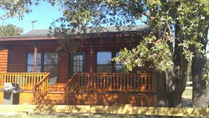 Una cabaña de troncos con una terraza de madera, una parrilla y un árbol en primer plano.