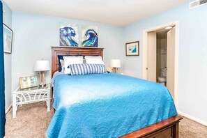 Un dormitorio con cama, mesitas de noche, una puerta y una pared con dos cuadros.