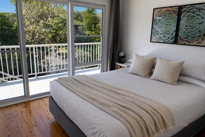 A bedroom with a large bed, a balcony with a white railing, and a view of trees and a building.