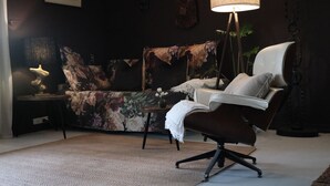 Een moderne woonkamer met een donkere muur, een bloemenbank, een bruine leren fauteuil, een klein bijzettafeltje met een lamp, en een vloerlamp.