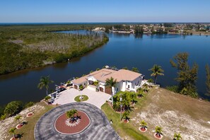 A large house with a spacious patio and a circular driveway, surrounded by a lake and palm trees.