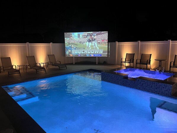 A swimming pool with a built-in hot tub and a large screen displaying a sports event.