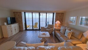 A spacious living room with a large sofa, a coffee table, and a television on a cabinet. There is a balcony with yellow chairs and a view of the ocean. - Sanibel Harbour Resort: Bay View Tower 1035 Relaxation awaits you at this beachfront retreat (Fort Myers)