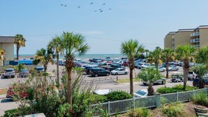 A parking lot with multiple cars, palm trees, and a building in the background. - Pavilion Watch 2B - Oceanview Condo - Pool - Central Location - Elevator (Folly Beach)