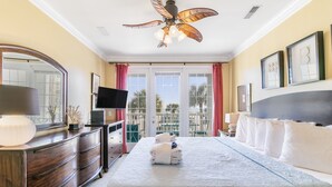A hotel room with a large bed, a wooden dresser, a flat-screen TV, a ceiling fan, and a balcony with a view of palm trees. - Pavilion Watch 2B - Oceanview Condo - Pool - Central Location - Elevator (Folly Beach)