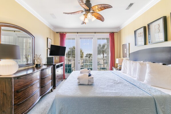 Habitación de hotel con una cama grande, cómoda de madera, televisor de pantalla plana, ventilador de techo y balcón con vista a palmeras.
