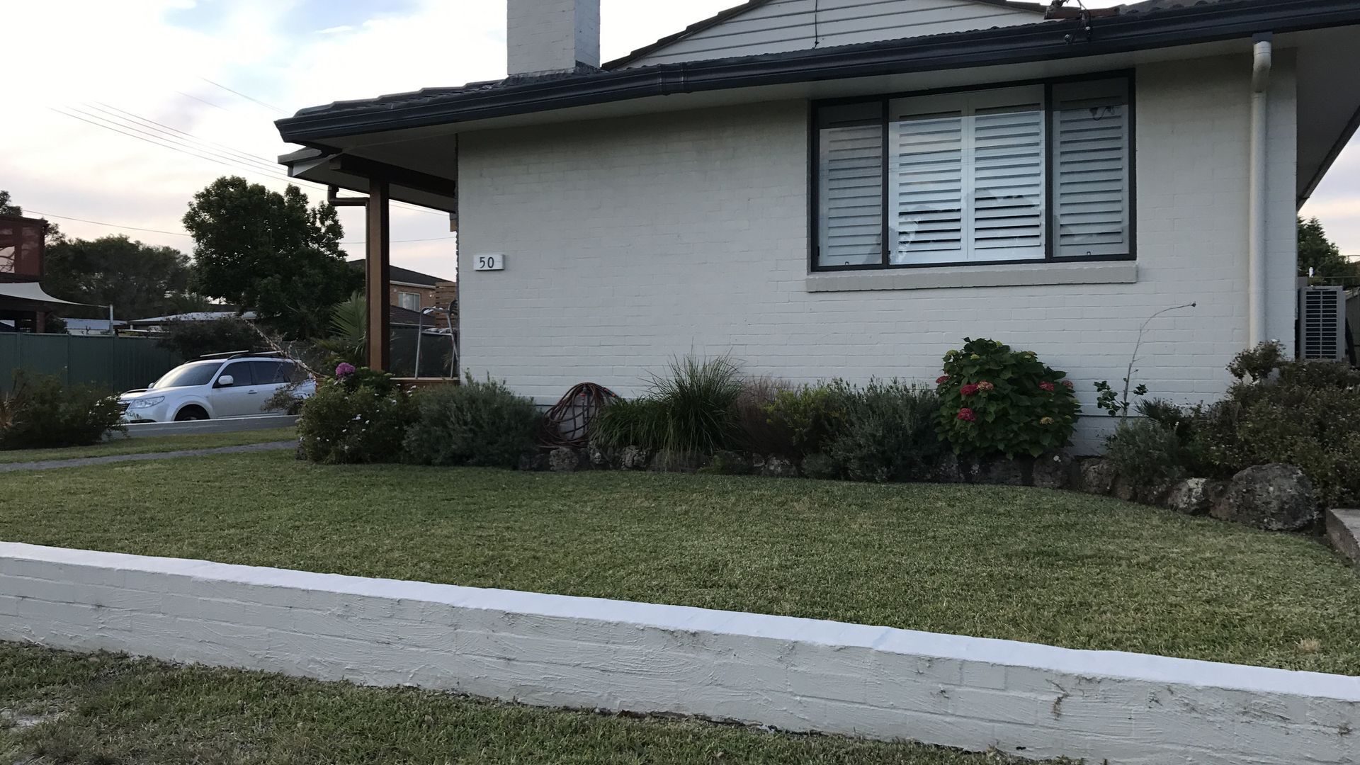 Uma casa térrea com fachada branca, o número '50' visível na lateral e um pequeno jardim frontal com grama e arbustos.
