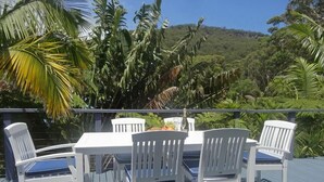 A deck with a table and chairs, surrounded by tropical foliage and a view of a hill. - Jacaranda House where beach meets rainforest (Stanwell Park)