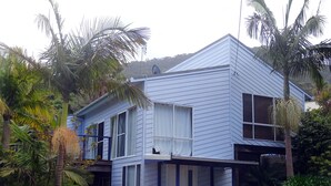 A two-story house with a blue exterior, a front porch, and a small garden with tropical plants. - Jacaranda House where beach meets rainforest (Stanwell Park)