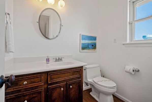 A bathroom with a white sink, wooden vanity, a round mirror, a toilet, and a painting on the wall. - 4 Bdr., 2.5 Bath Beachfront Home. Recently painted and a fresh kitchen remodel. (Galveston)
