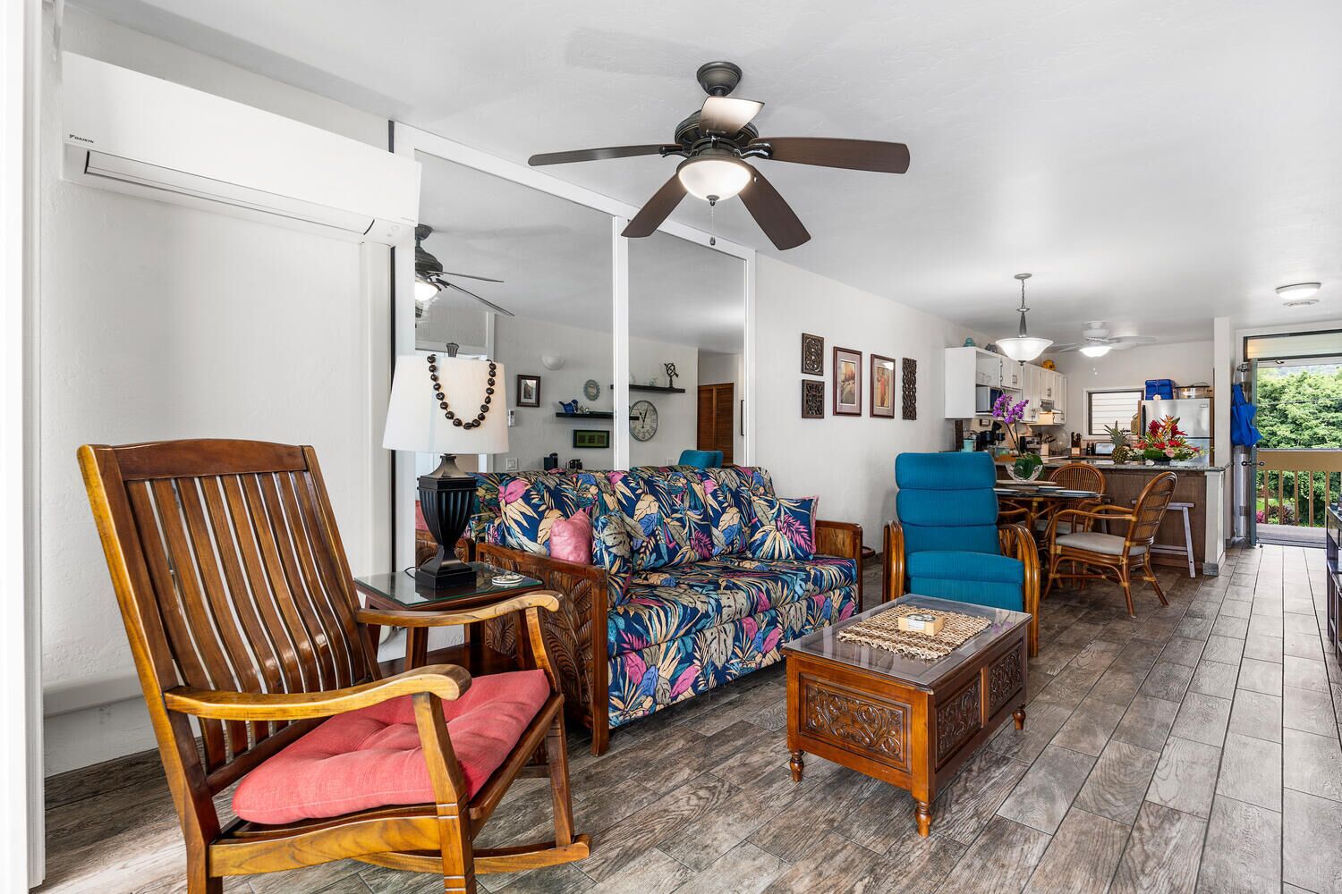 Ein geräumiges Wohnzimmer mit Deckenventilator, einer floralen Couch, einem Essbereich mit Tisch und Stühlen sowie einer durch einen offenen Durchgang erkennbaren Küche.