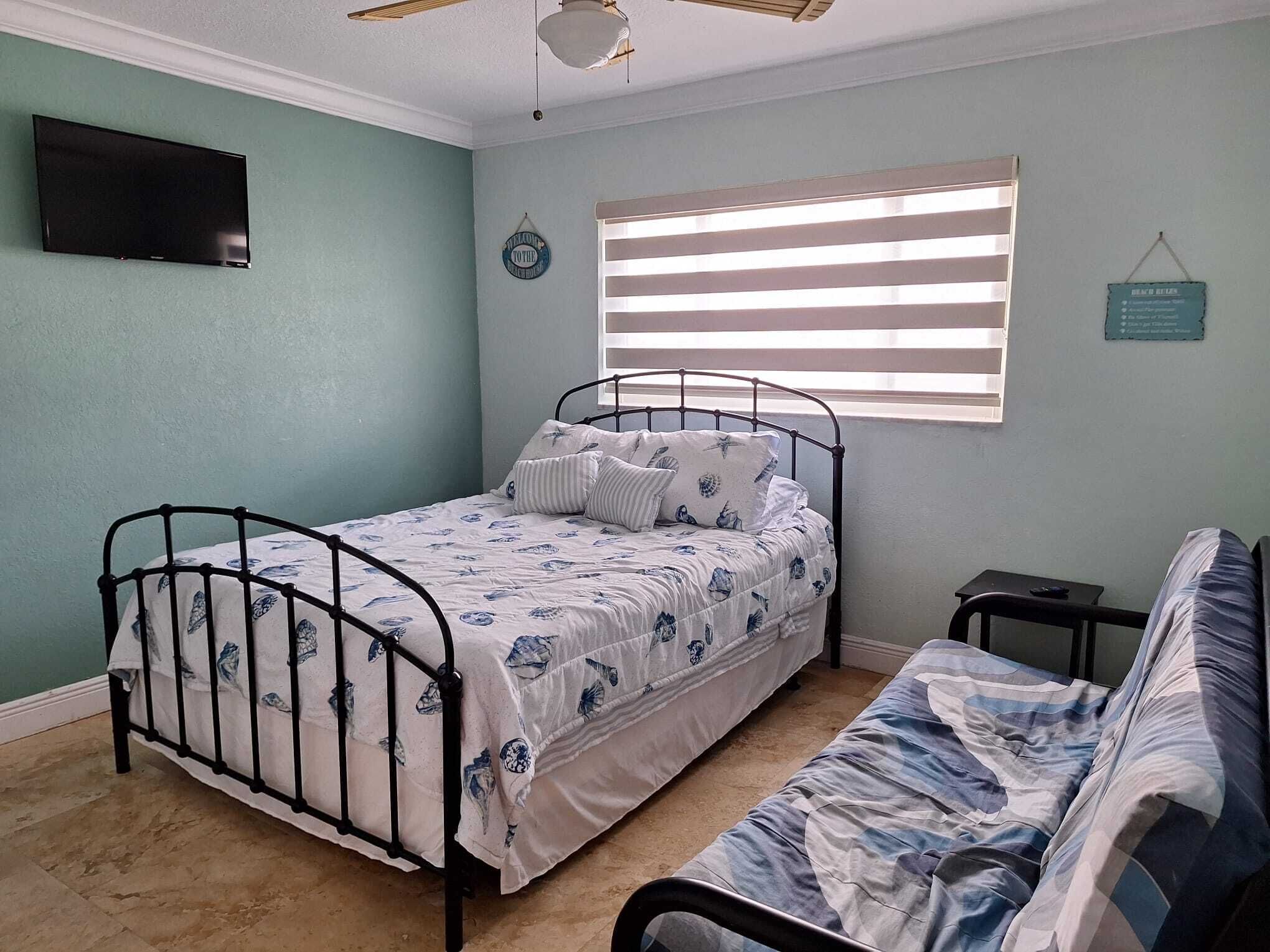 A bedroom with a bed, a bench, a TV, and a window with blinds.