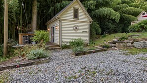 A small, single-story cabin with a sloped roof, surrounded by dense greenery and a gravel driveway. - Beautiful Waterside Cabin (2), Great Location, View, 8 Min To Downtown, Biltmore (Asheville)
