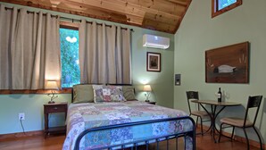 A bedroom with a bed, bedside tables, a window with curtains, a table with chairs, and a painting on the wall. - Beautiful Waterside Cabin (2), Great Location, View, 8 Min To Downtown, Biltmore (Asheville)