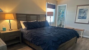 A bedroom with a wooden bed, a nightstand with a lamp, a bedside table, and a chair. - BEACHFRONT LARGE SOUTHEND 3 BEDROOM 2 BATH CONDO (Brigantine)