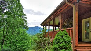 A wooden cabin with a porch, surrounded by lush greenery and mountains in the background. - Experience comfort at Incommunicado in Warrensville, NC—a cozy 4-bedroom cabin for 10! (Warrensville)