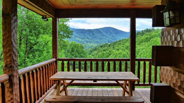 A wooden deck with a picnic table, stone wall, and a view of a forested mountain range. - Experience comfort at Incommunicado in Warrensville, NC—a cozy 4-bedroom cabin for 10! (Warrensville)