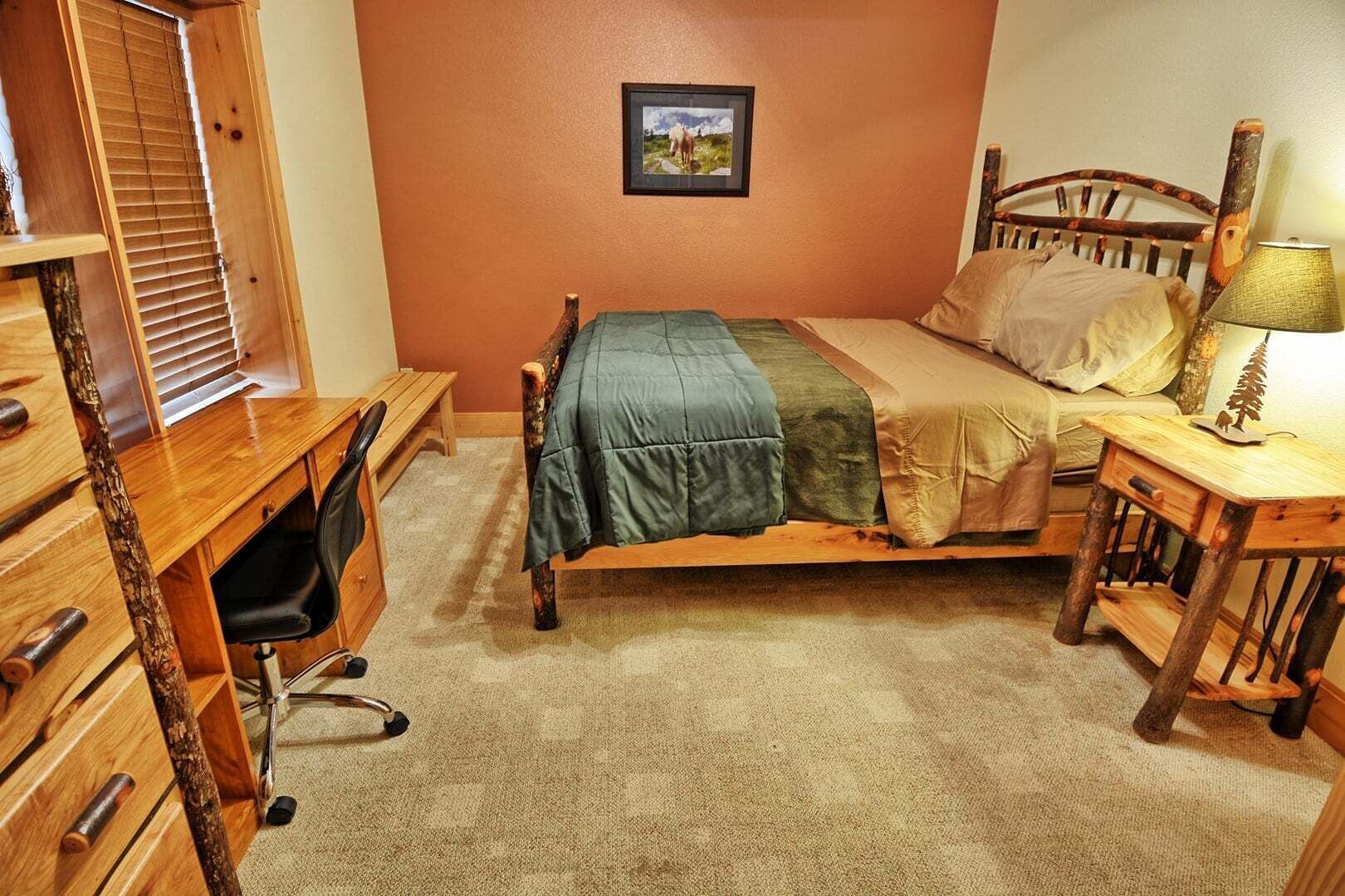 A bedroom with a wooden desk, a bed with a green blanket, a wooden nightstand with a lamp, and a picture on the wall.