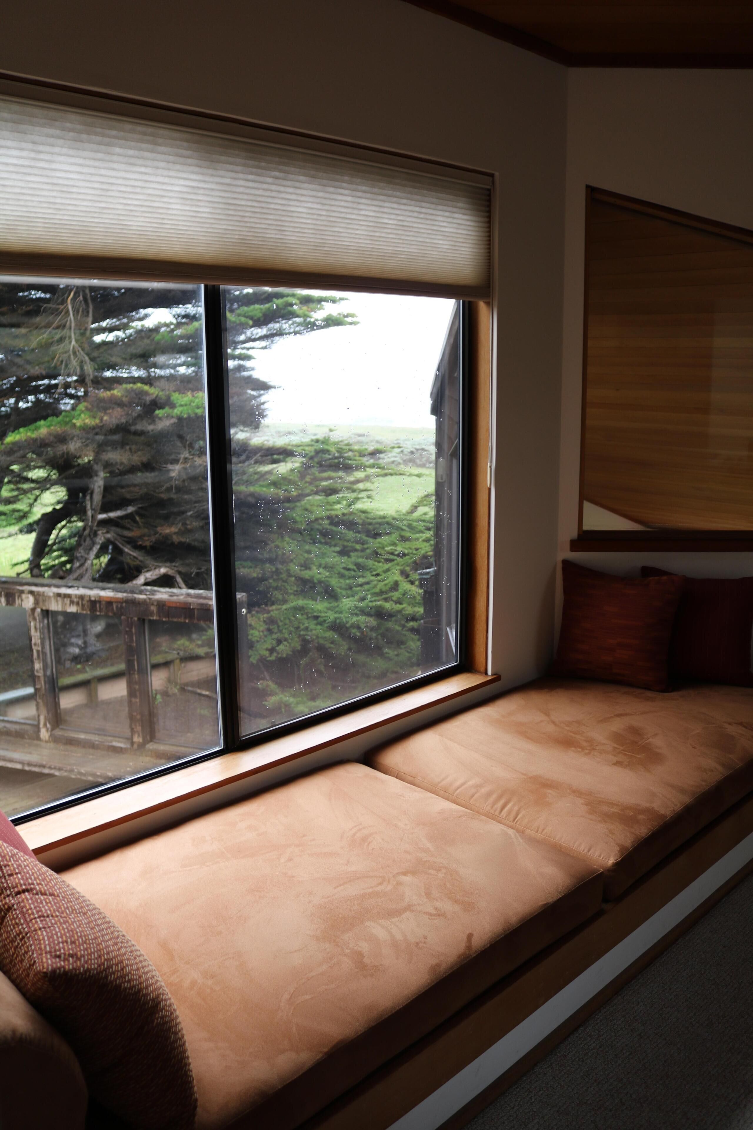 A room with a large window, a cushioned bench, and a view of a forest.