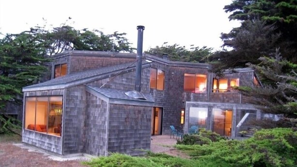 A modern house with large windows and a chimney, surrounded by greenery. - OCEAN FRONT-WHITE WATER VU from every room - above Del Mar Cove (The Sea Ranch)