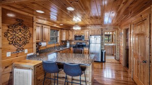 A wooden cabin interior with a kitchen featuring granite countertops, a central island with bar stools, and a built-in stove.