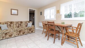 Un salon avec un canapé à motifs floraux, une table à manger en bois et des chaises. On y trouve une porte qui mène à une autre pièce et une fenêtre ornée de rideaux en dentelle.