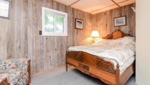 Une chambre avec un mur en bois, un lit recouvert d’une couvre-lit à motifs floraux, une table de chevet avec une lampe et une chaise agrémentée d’un coussin à motifs floraux.