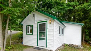 Une petite cabane à un seul étage, avec un toit vert et un revêtement blanc, entourée d’arbres.
