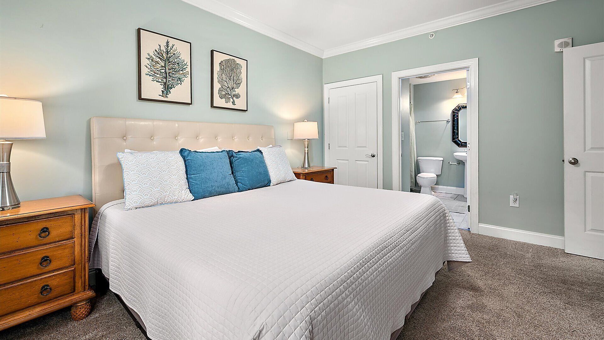 Quarto com cama grande, duas luminárias de cabeceira, mesa de cabeceira e cômoda. Há duas gravuras botânicas emolduradas na parede acima da cama.