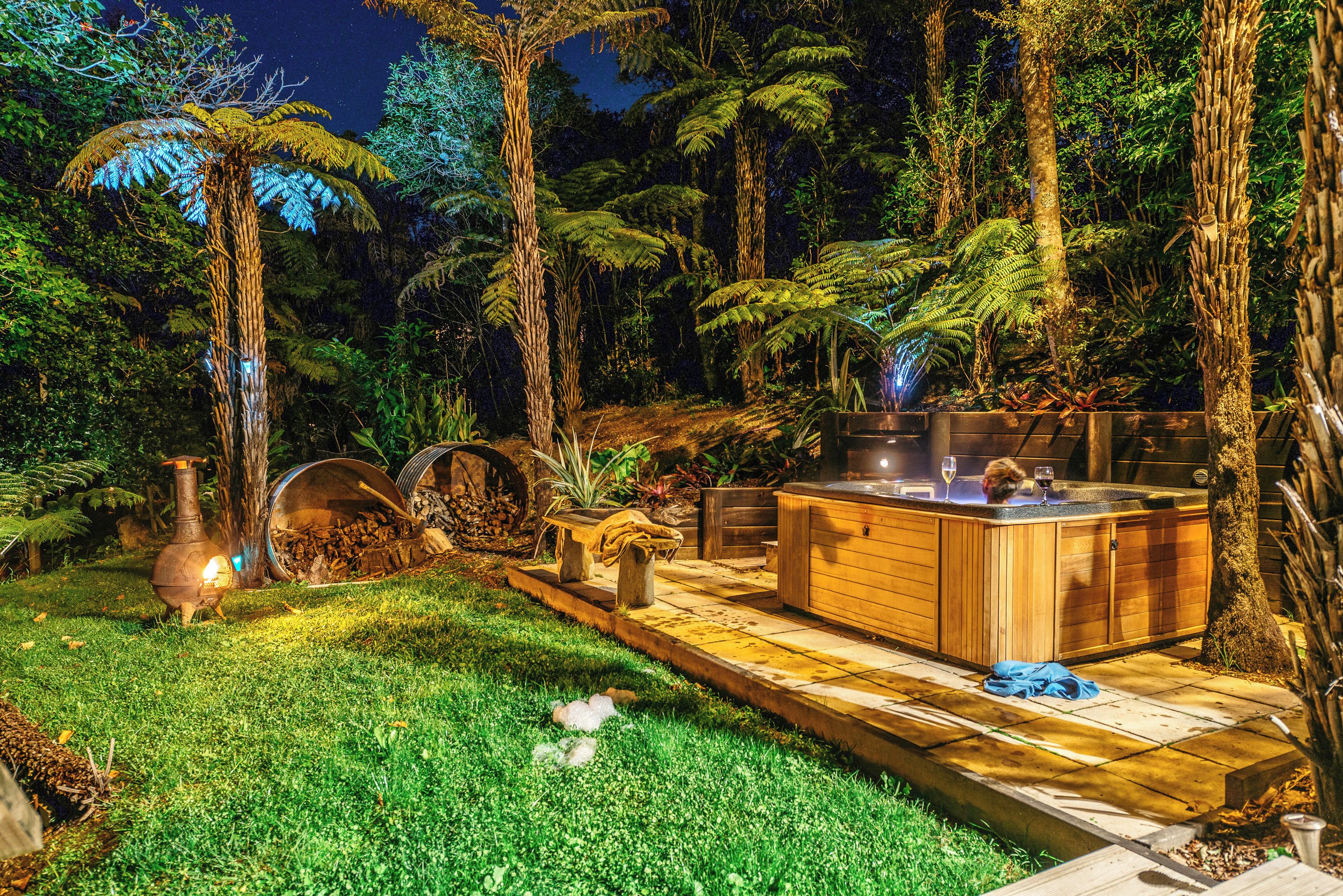 A well-lit outdoor area with a hot tub, wooden deck, and lush greenery including ferns and trees.