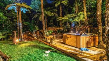A well-lit outdoor area with a hot tub, wooden deck, and lush greenery including ferns and trees.