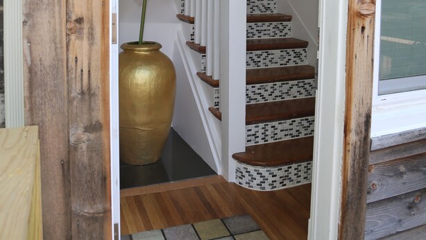 Puerta de madera que da a una escalera con barandilla blanca y base de mosaicos. Un jarrón dorado de gran tamaño con una flor blanca está colocado en los escalones.