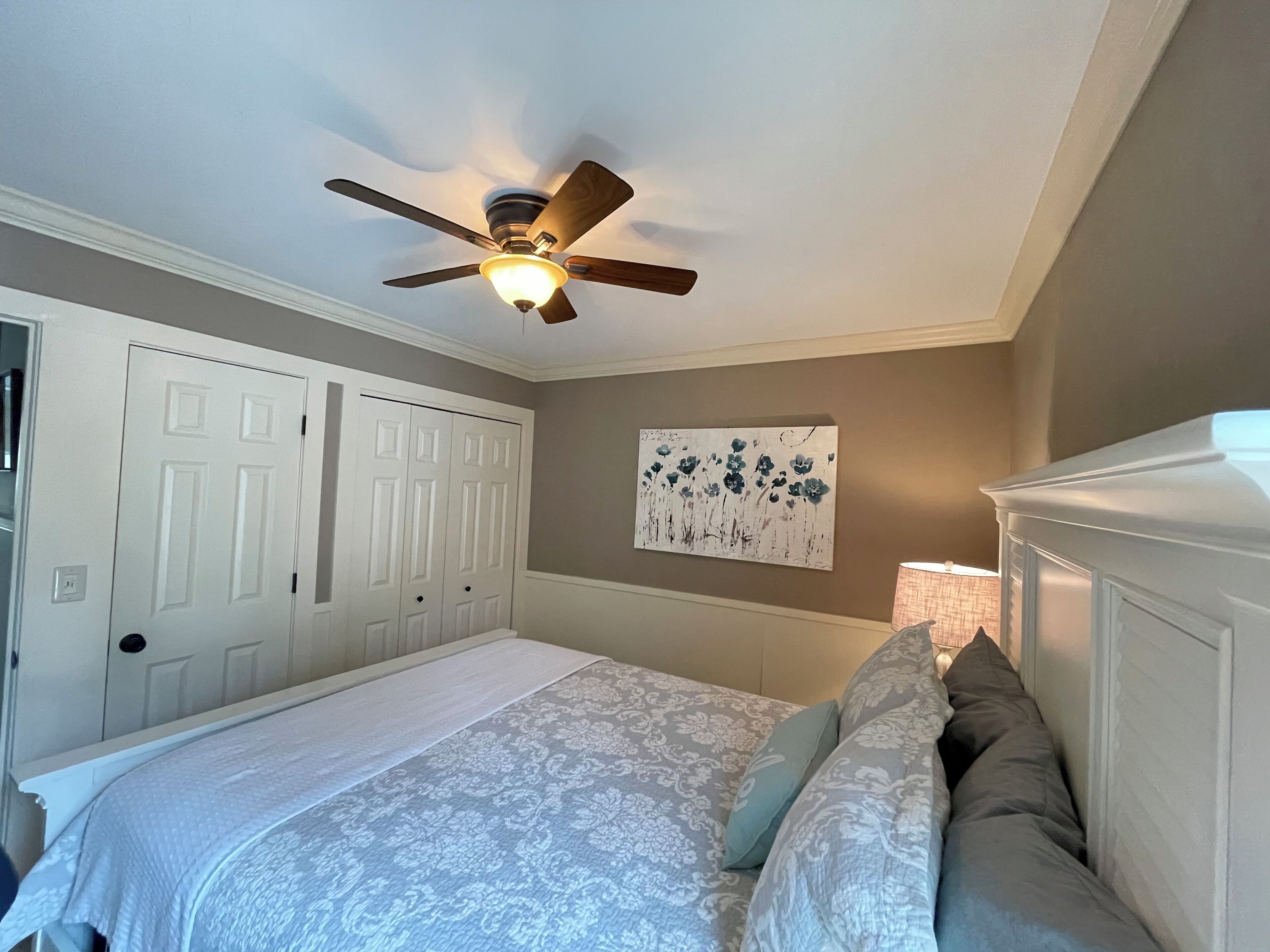 A bedroom with a bed, a ceiling fan, a door, and a wall-mounted lamp.