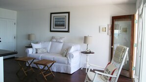 Una sala de estar blanca con un sofá, dos mesitas de madera, una silla y un cuadro en la pared.