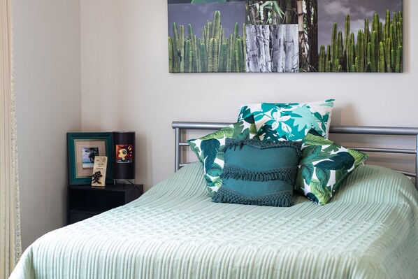 A neatly made bed with a green bedspread and multiple pillows, a framed picture on a nightstand, and a cactus-themed artwork on the wall.