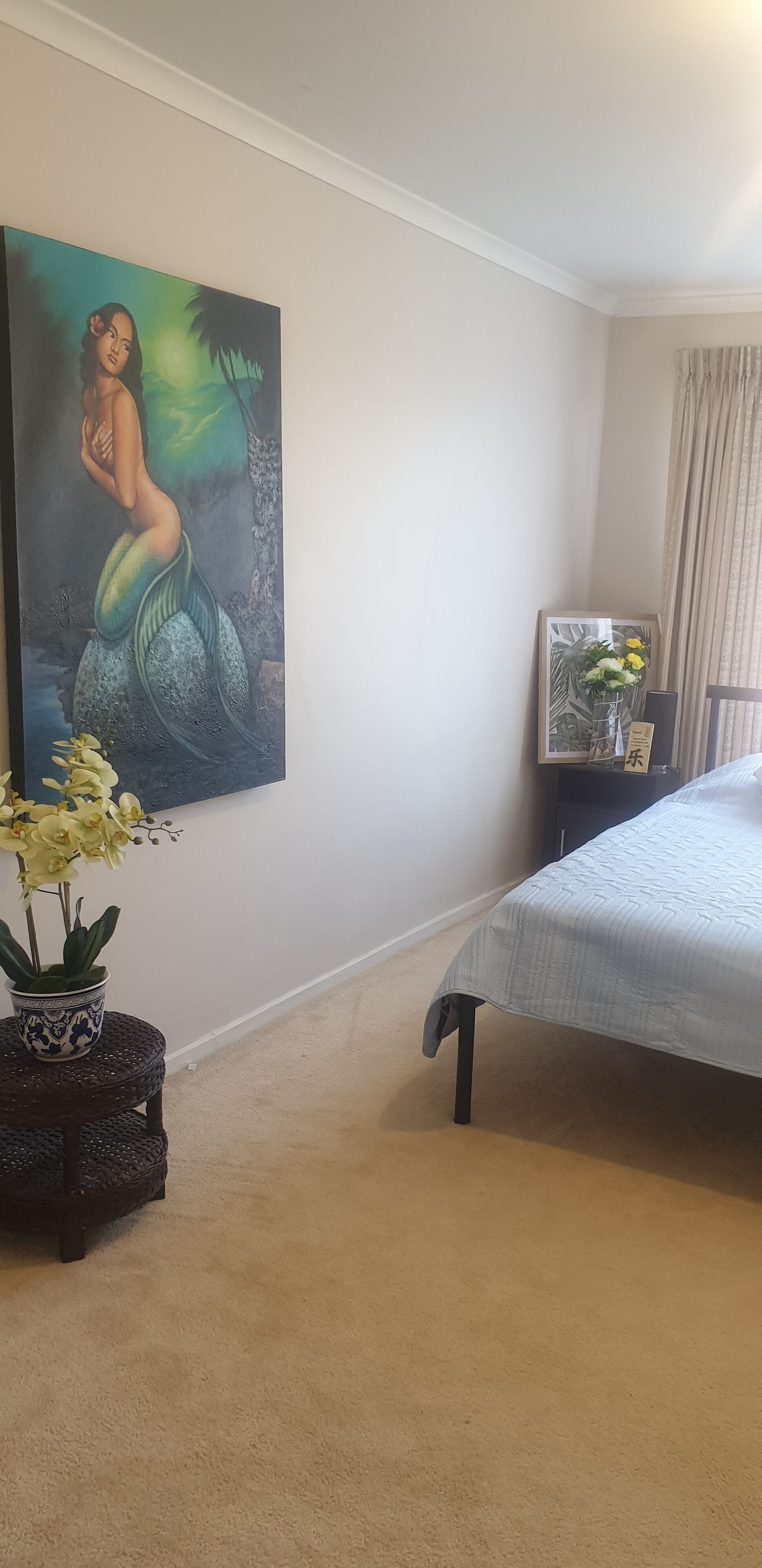 Een slaapkamer met een bed, een schilderij aan de muur, een klein tafeltje met een bloevas en een nachtkastje met een lamp.