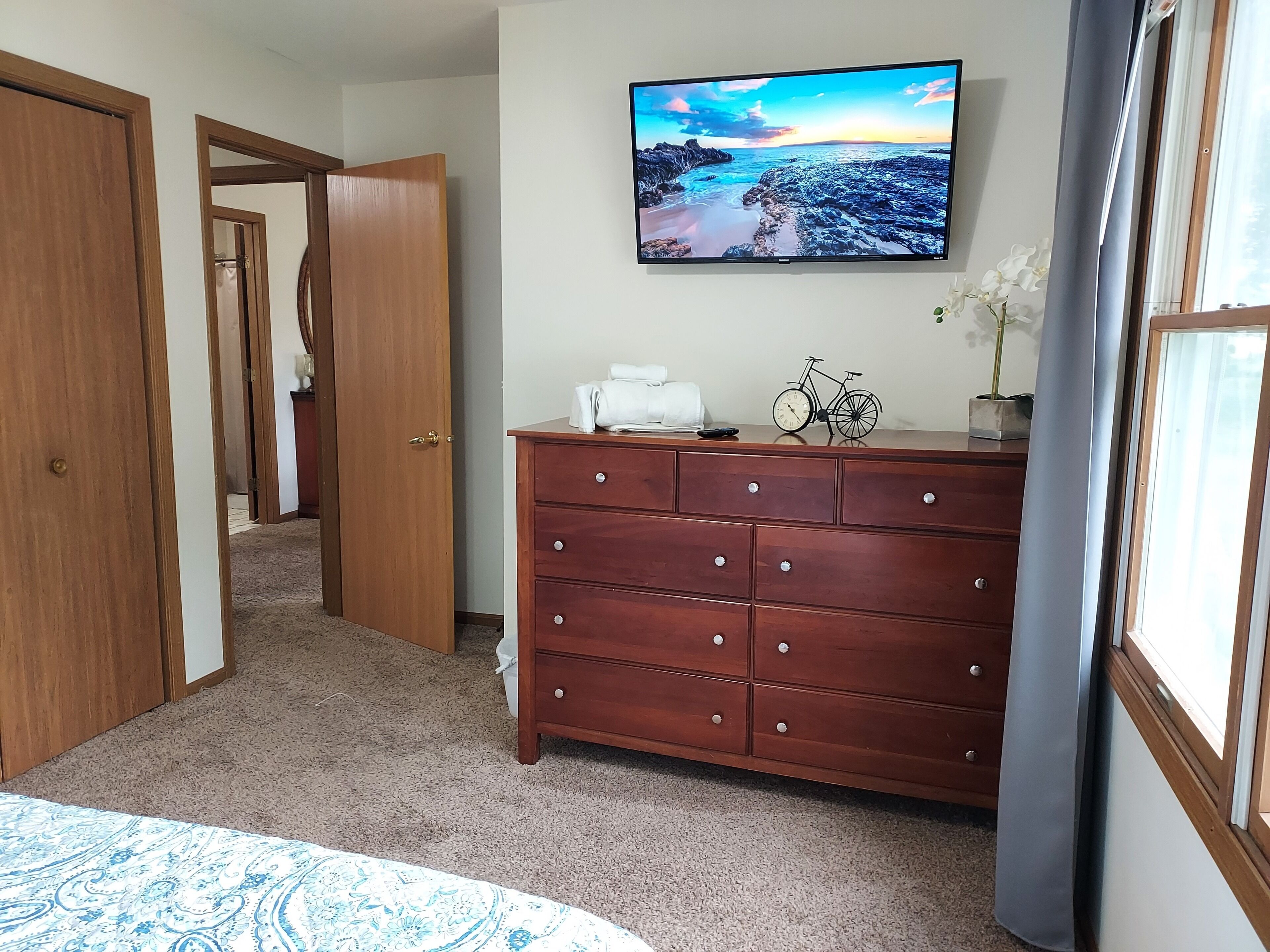 Une chambre à coucher comprenant un lit, une commode, un téléviseur diffusant un paysage pittoresque, et une porte donnant sur une autre pièce.