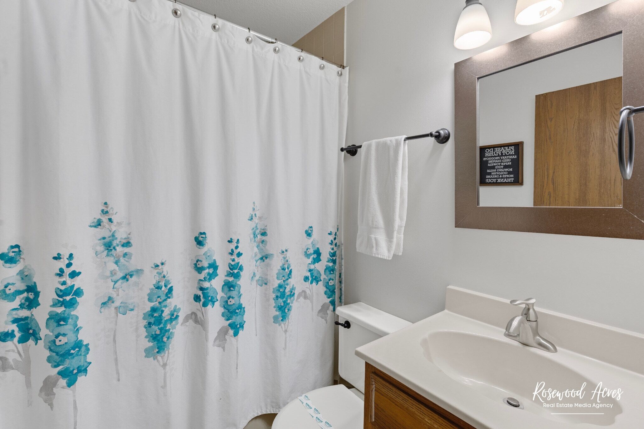 A bathroom with a white shower curtain featuring blue floral patterns, a white toilet, a sink with a mirror above it, and a sign on the wall.