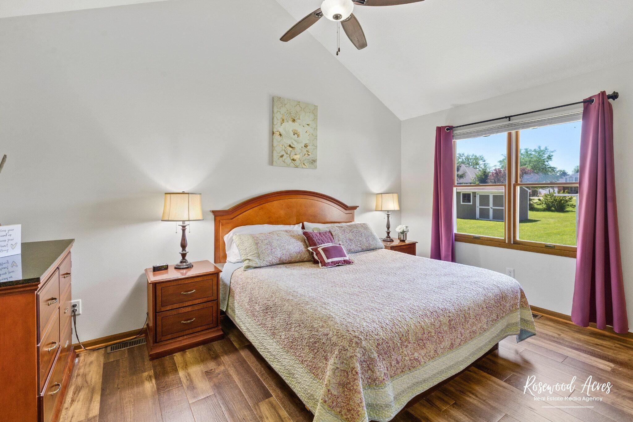 Une chambre à coucher avec un lit en bois, deux lampes de chevet, un ventilateur de plafond, une fenêtre avec des rideaux et une vue sur l’extérieur.
