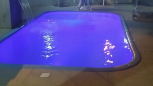 Piscina iluminada con luz azul, rodeada de sillas y una mesa blancas bajo una pérgola.
