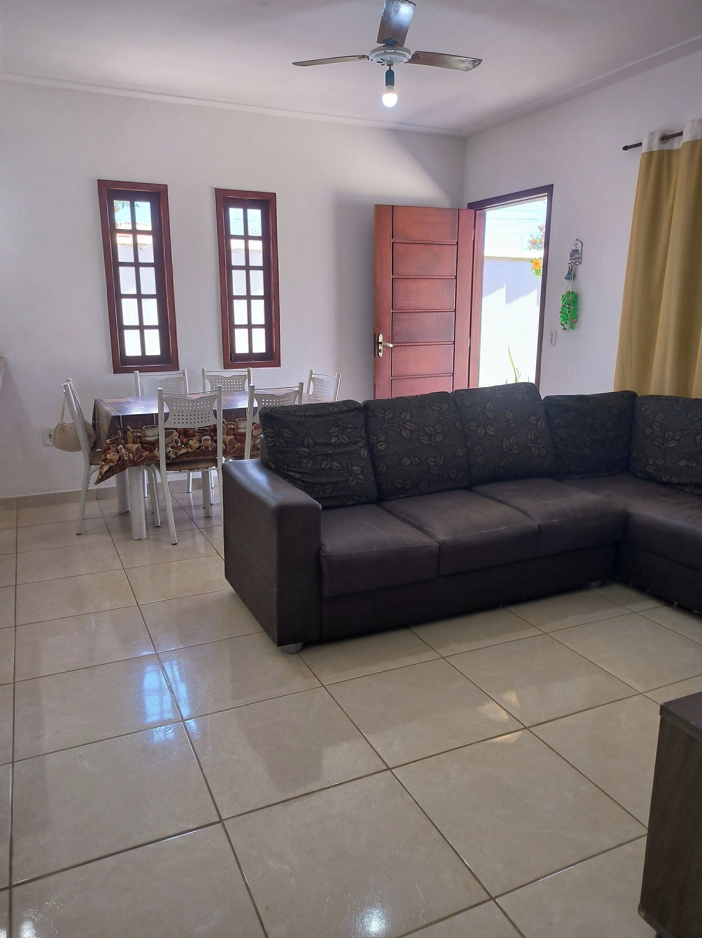 A living room with a dark brown sofa, a dining table with chairs, two windows, a ceiling fan, and a door.