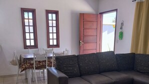Una sala de estar con un sofá marrón oscuro, una mesa de comedor con sillas, dos ventanas, un ventilador de techo y una puerta.