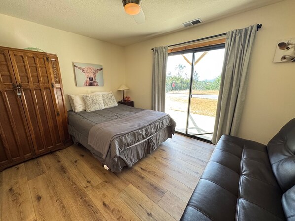 A bedroom with a wooden wardrobe, a bed with grey bedding, a nightstand with a lamp, and a leather sofa.
