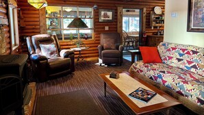 Un accogliente interno di una baita in legno con una poltrona in pelle, un divano con una coperta colorata, un tavolino in legno e un camino.